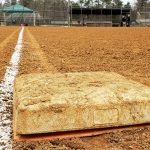 Dirty first base on a baseball field looking toward home and third base dugout. Bright white foul line on clay diamond. Low angle. Little league baseball field in public park.